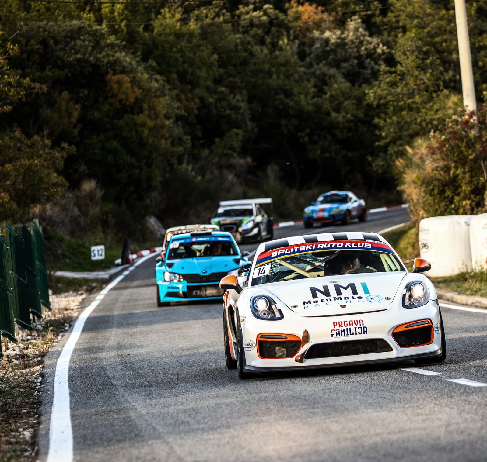 A white Porsche Cayman leads three cars up the road between the trees
