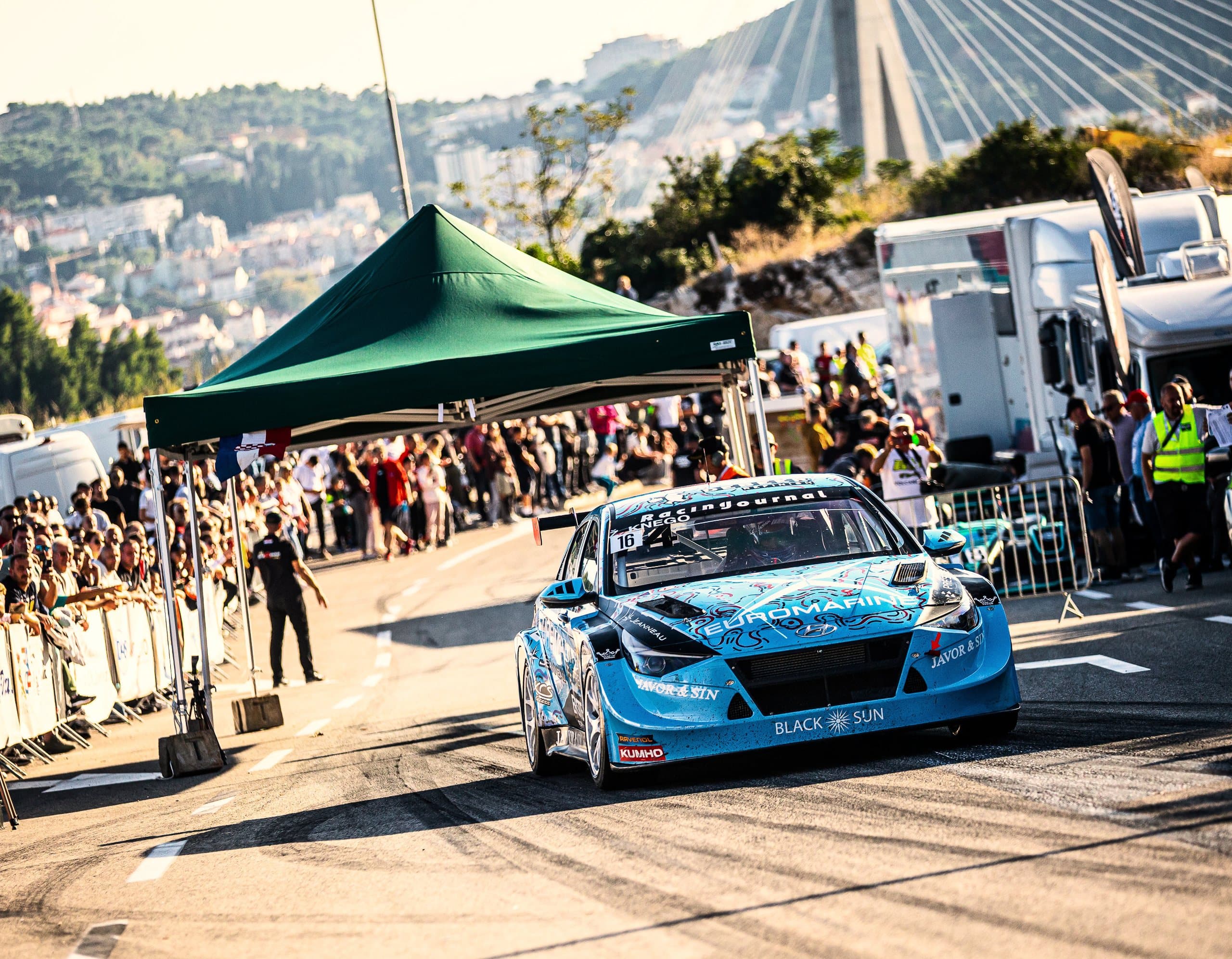 Žarko Knego's light blue Hyundai no. 16 at the start under the bridge, spectators along the barriers