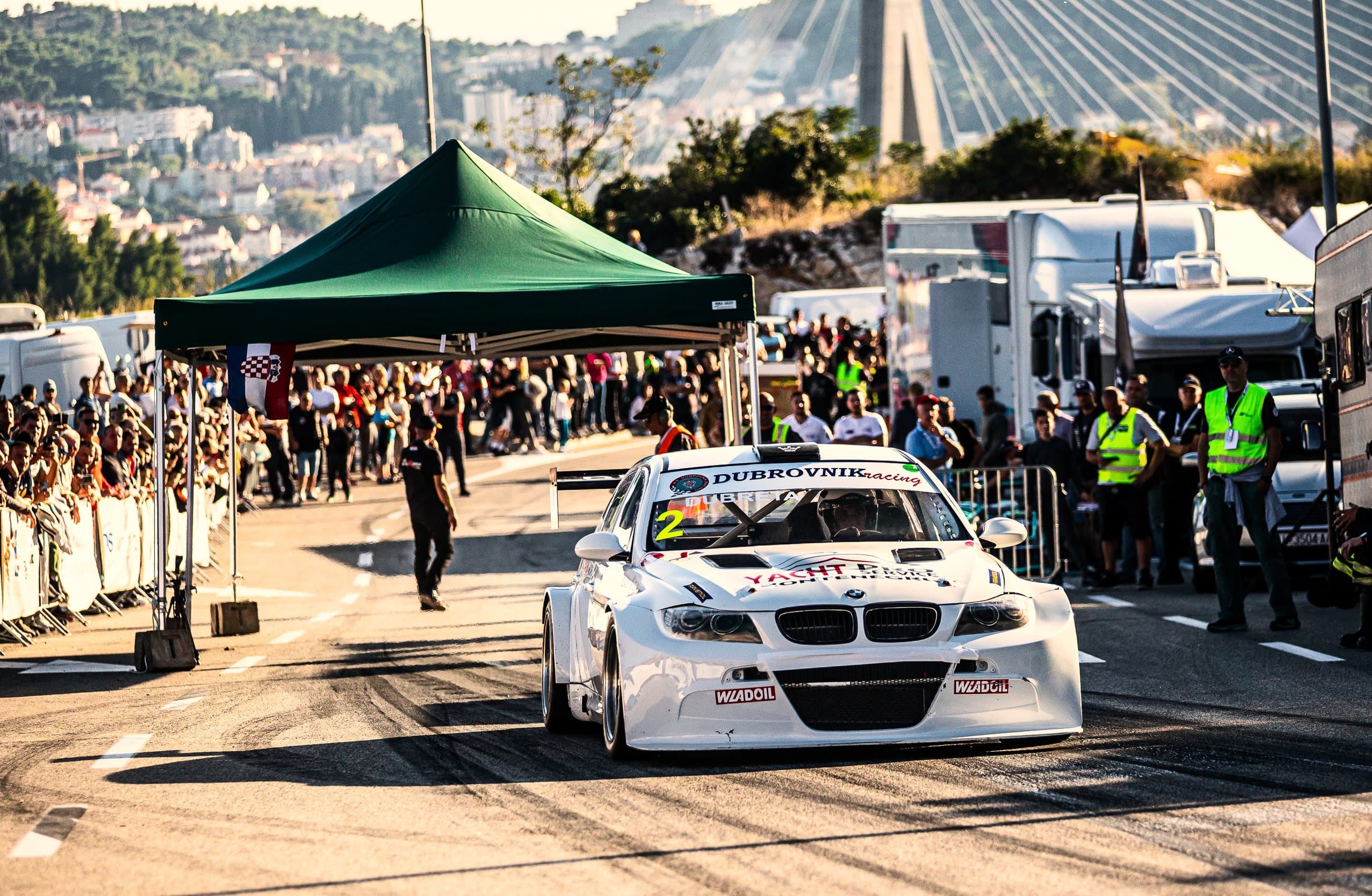 The white BMW leaves the start under the bridge, spectators along the barriers